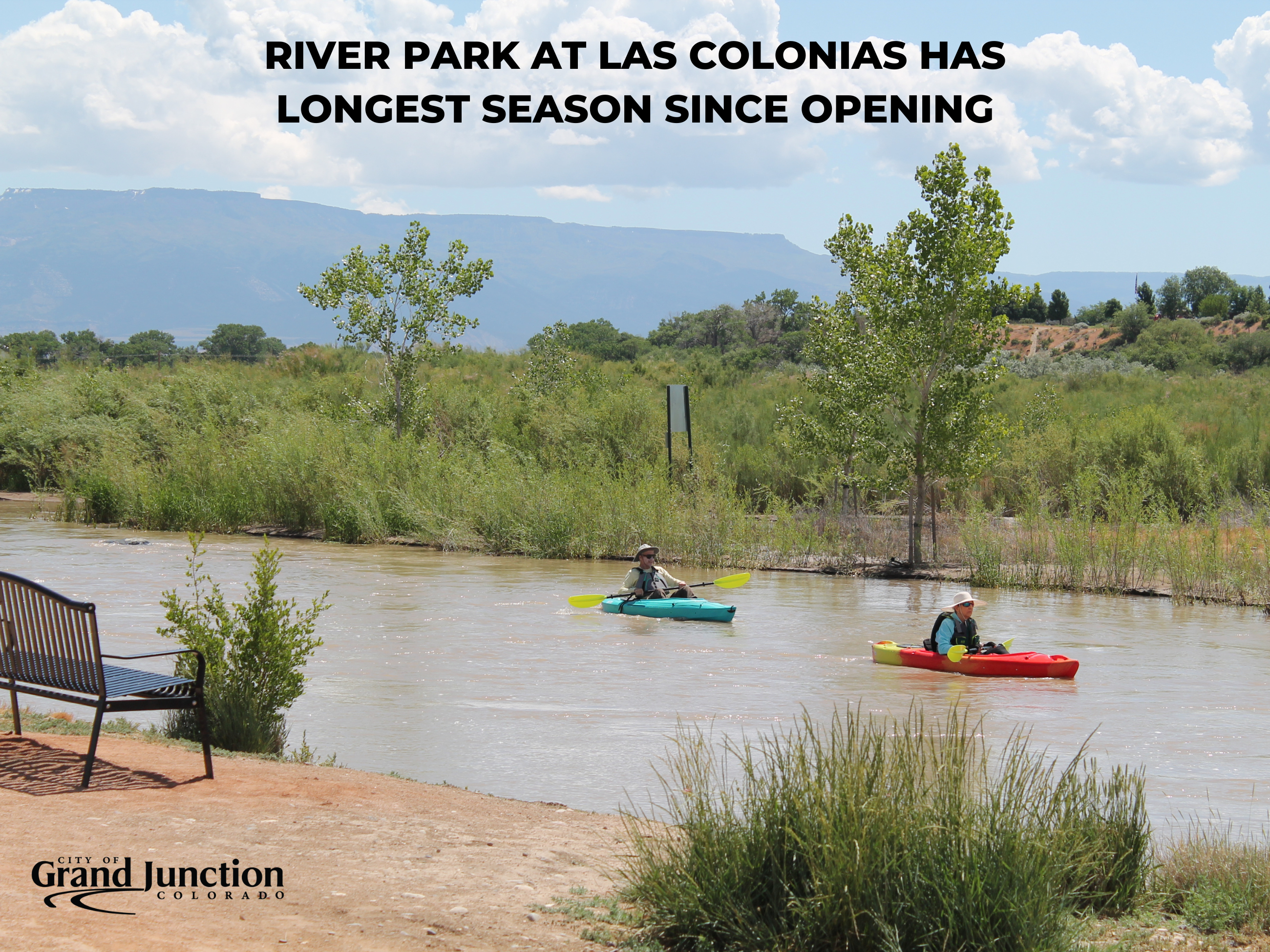 Shoreline with bench and two people in kayaks in river. 