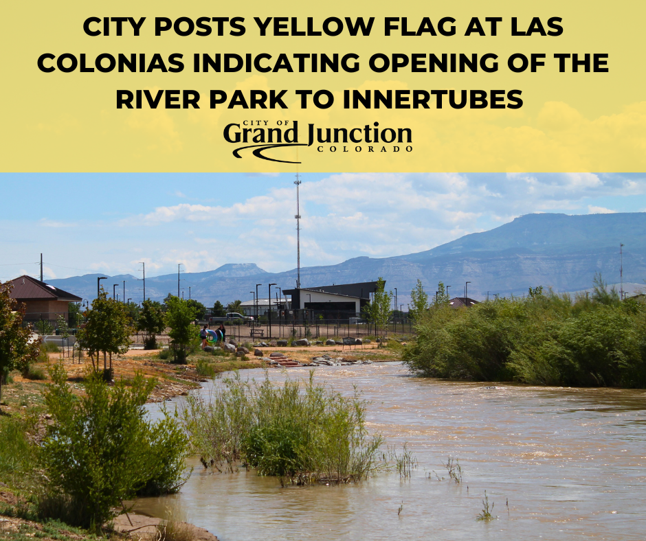 A shallow river flowing through brushes. Buildings in the background with people.
